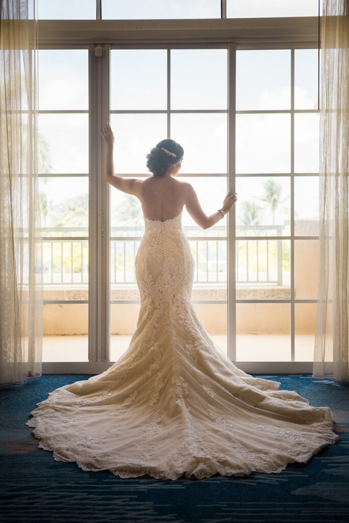 Bride in wedding dress standing by window with back to camera