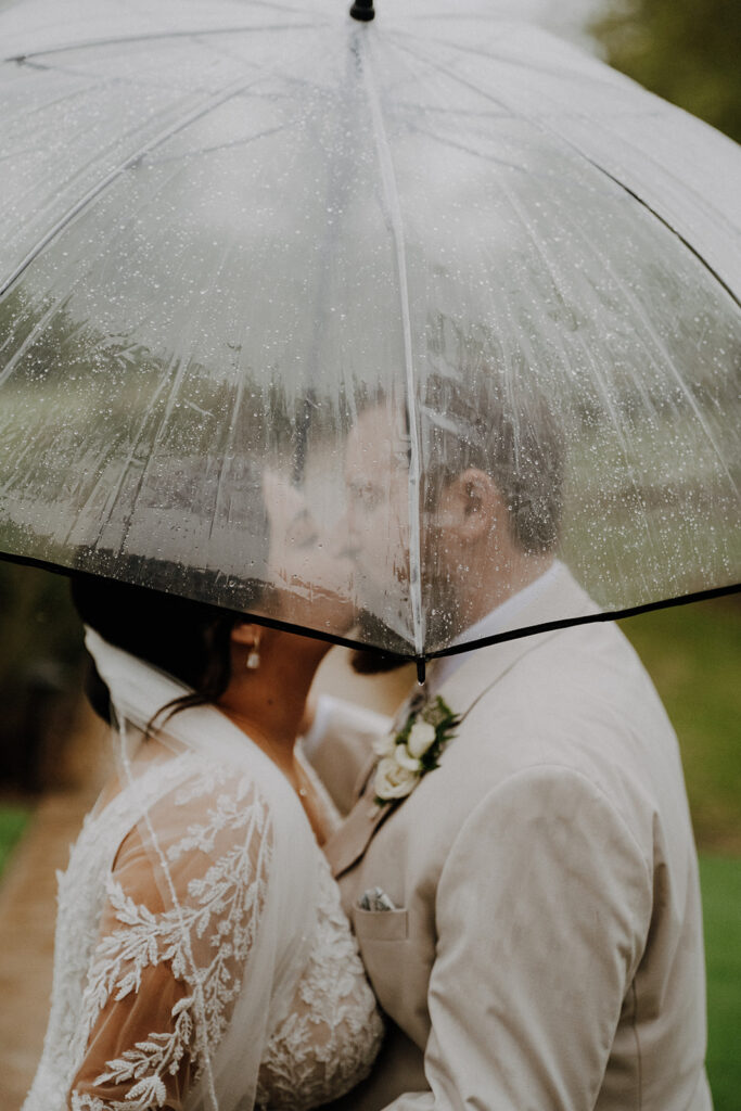 bride and groom caught kissing under an umbrella while it rains during the wedding