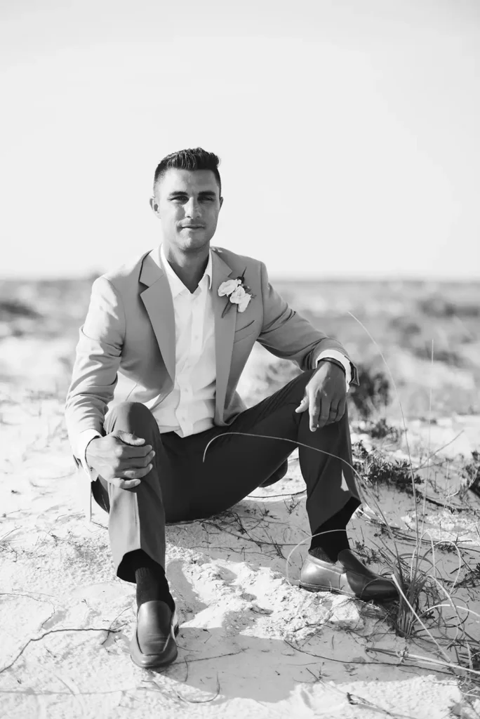 Black and white photo of groom sitting on sandy beach