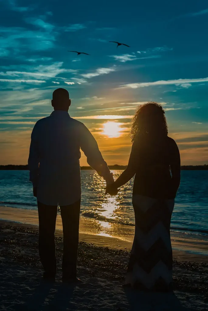 Man and woman holding hands by ocean with sun setting