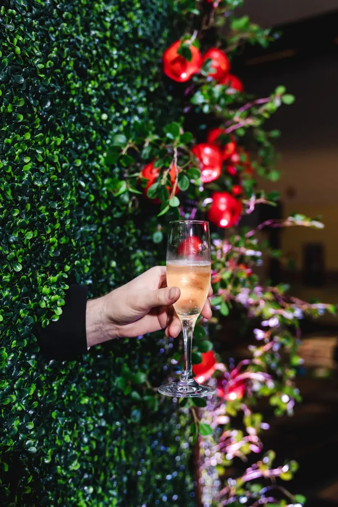 Hand sticking through greenery wall holding glass of champagne at corporate event