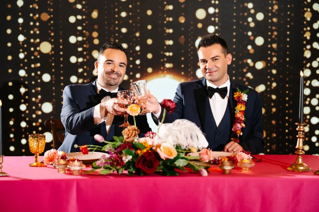 Two grooms clink glasses while sitting at head table